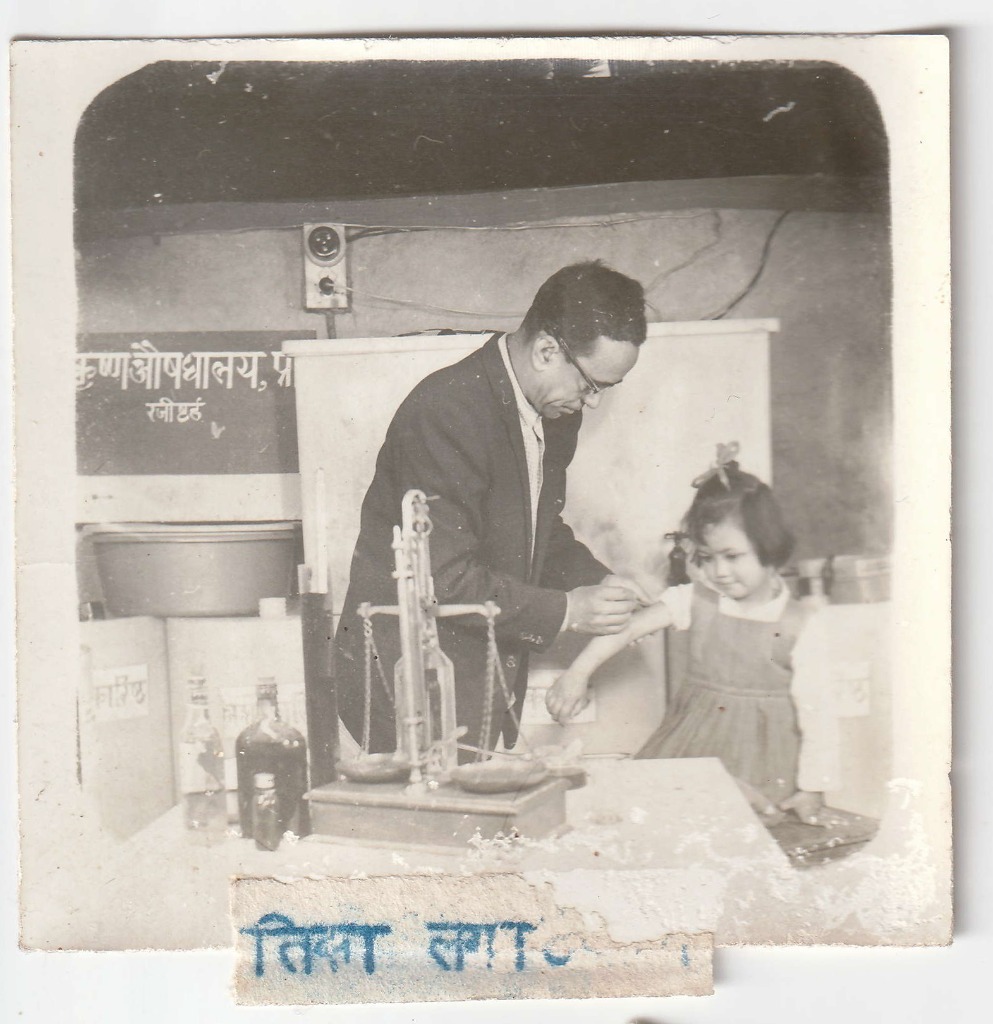 A senior member of the Baidya family weighing medicine on an apothecary balance with a child watching. Date unrecorded.
