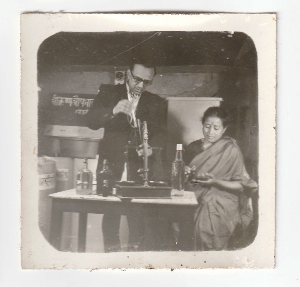 Two members of the Baidya family at the Shree Krishna Aushadhalaya dispensary counter with balance scale and registered signboard. Date unrecorded.