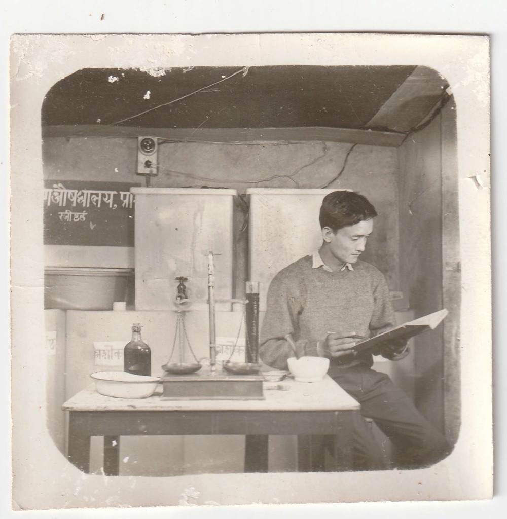 A member of staff reading a formulary record at the Shree Krishna Aushadhalaya dispensary workbench. Date unrecorded.